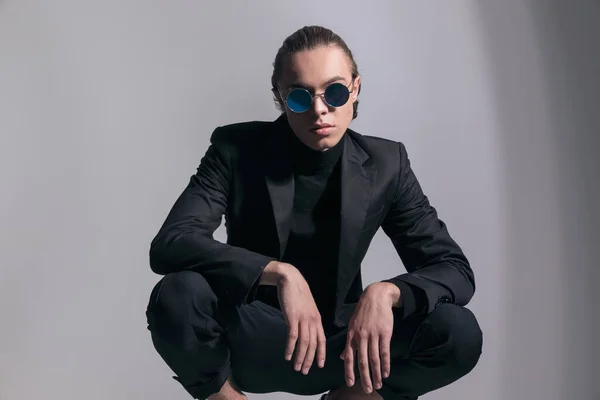 Fashion picture of young businessman squatting and posing with cool vibe and wearing a nice outfit against gray studio background