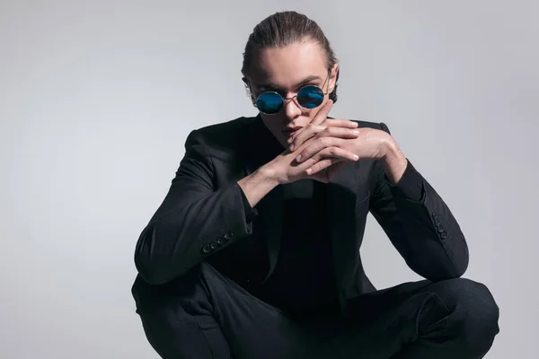Fashion picture of sexy businessman crossing his fingers in squatted position and wearing a nice outfit against gray studio background