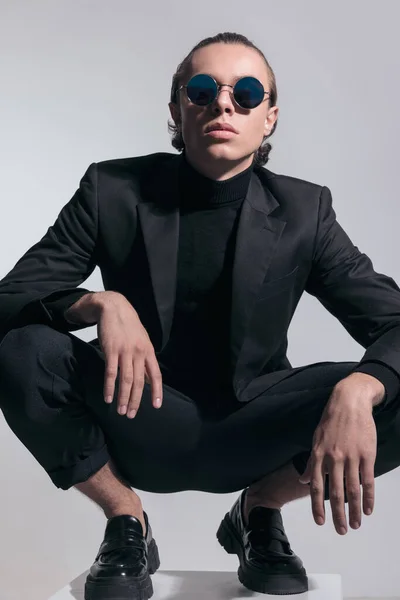 Fashion picture of young businessman squatting like a boss and wearing a nice outfit against gray studio background