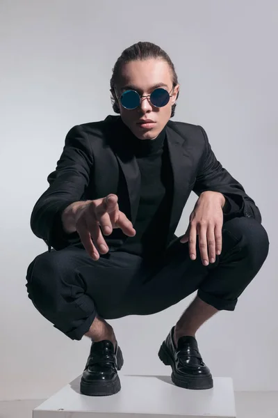 Fashion picture of attractive businessman squatting and pointing at camera and wearing a nice outfit against gray studio background