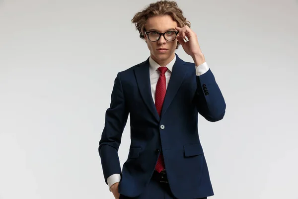 有魅力的商人手插在口袋里 修理眼镜 在灰色工作室背景下穿上正式服装的肖像 — 图库照片