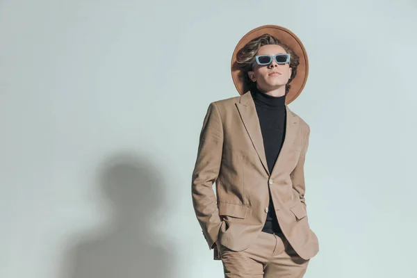 stock image cool young man with hat and sunglasses holding hands in pockets and posing in front of grey background