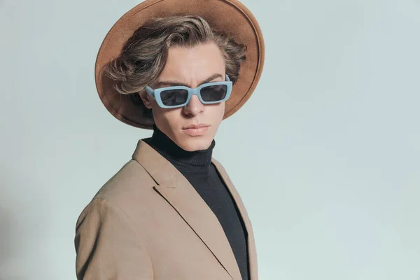 portrait of cool fashion guy with long hair wearing hat and sunglasses and posing in front of grey background