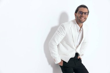 Gözlüklü, beyaz ceketli, yaka gömlekli, cebinde el ele tutuşan ve gri arka planda gülen çekici bir adam.