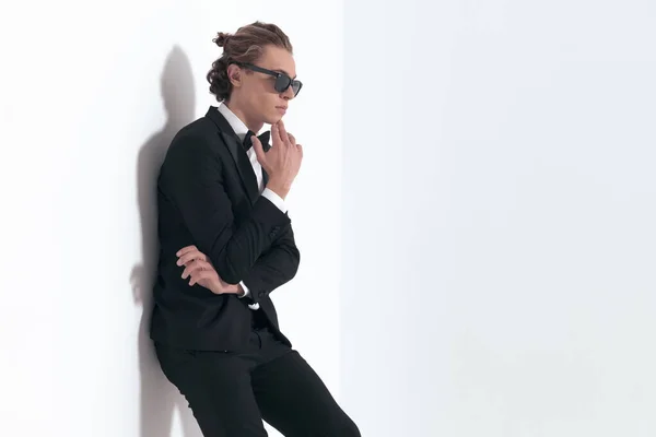 thoughtful elegant groom with sunglasses laying on a wall and thinking in front of grey background