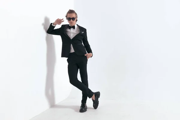 sexy young man in tuxedo with sunglasses laying his elbow on a grey wall, posing with hand in pockets in front of grey background