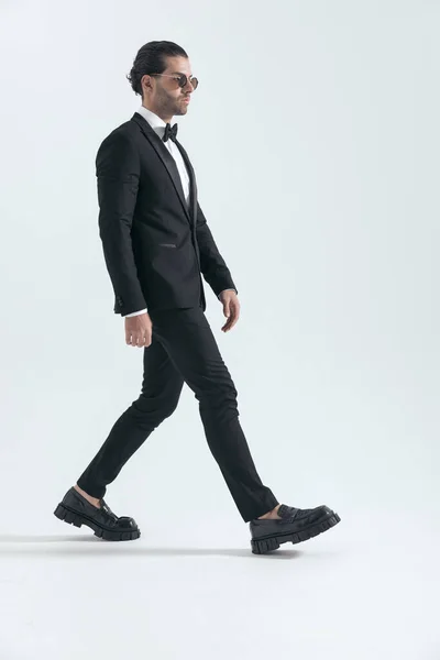 时尚优雅的男人 戴着墨镜 身穿黑色燕尾服 面朝一边 走在工作室灰色背景的前面 — 图库照片