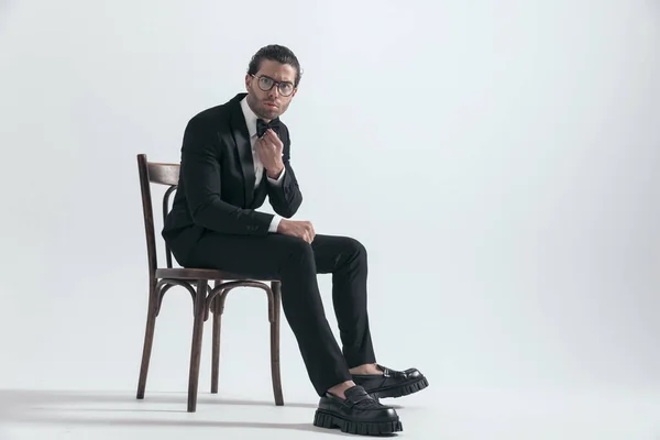 穿着黑色燕尾服的严肃而优雅的男人在灰色背景的木椅上皱着眉头固定领带 — 图库照片