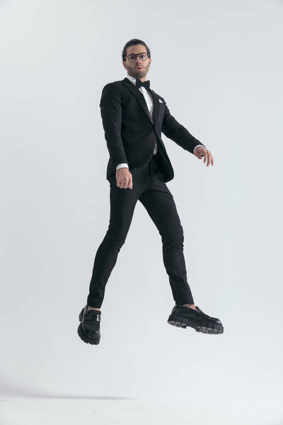 fashion young man in black tuxedo with glasses leapging up in the air on grey background