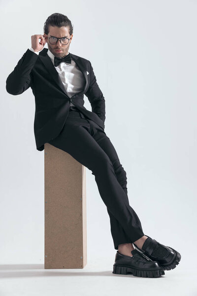 elegant young groom in black tuxedo fixing glasses and sitting with hand in pocket in front of grey background