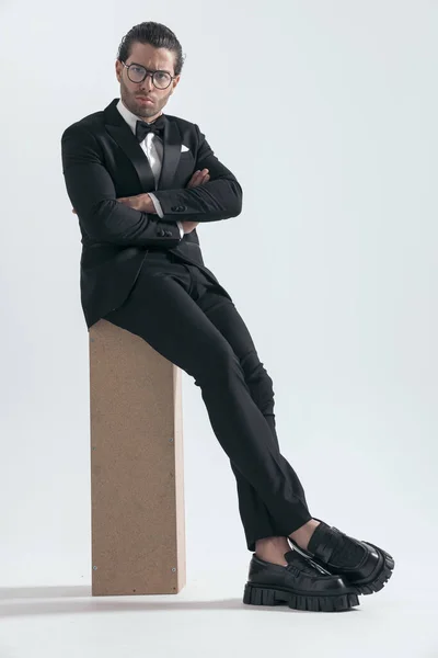 sexy fashion man in tuxedo crossing arms and sitting in a cool way on grey background