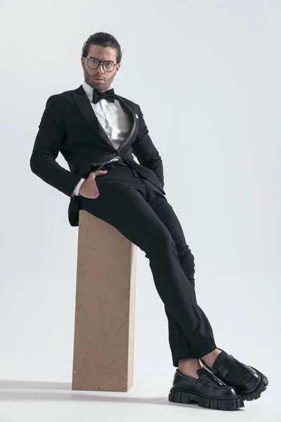 cool fashion man in tuxedo sitting with hands in pockets and posing in a confident way on grey background
