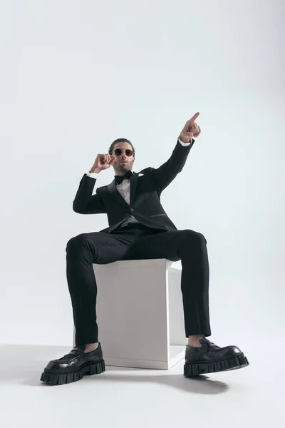 时尚优雅的男人 戴着太阳镜远眺 手指指向远方 坐在灰色的背景上 — 图库照片