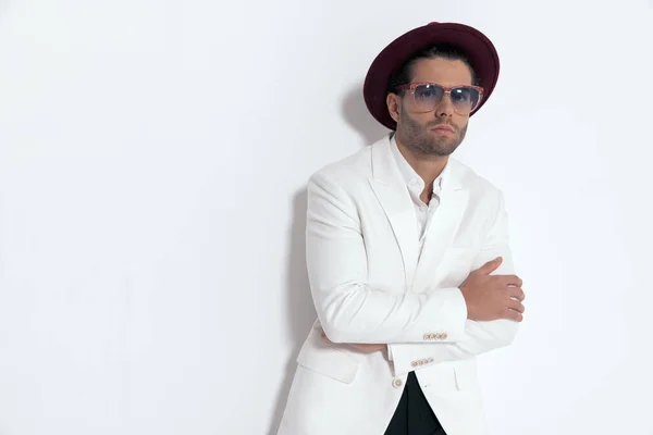 cool elegant man with hat and glasses crossing arms and posing while laying on a grey wall