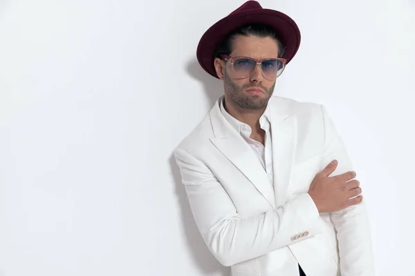 cool young man wearing hat and sunglasses holding arm in fashion way and posing in front of grey background