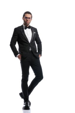 Yakışıklı iş adamı bacak bacak üstüne atıyor, elini cebine sokuyor ve beyaz stüdyo geçmişine karşı gözlük takıyor.