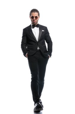 Zarif bir iş adamı, havalı yürüyor, elleri ceplerinde ve beyaz stüdyo geçmişine karşı gözlük takıyor.