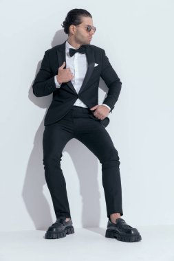 Seksi iş adamı duvara yaslanıyor ve smokinini ayarlıyor, beyaz stüdyo arka planına karşı gözlük takıyor.