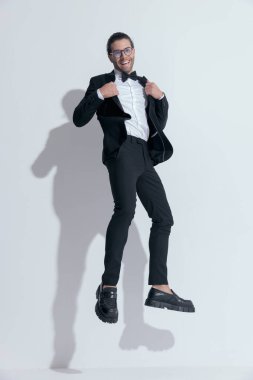 Çekici iş adamı smokinini düzeltirken ve havada gülerken yakalandı, beyaz stüdyo arka planında gözlük takıyordu.