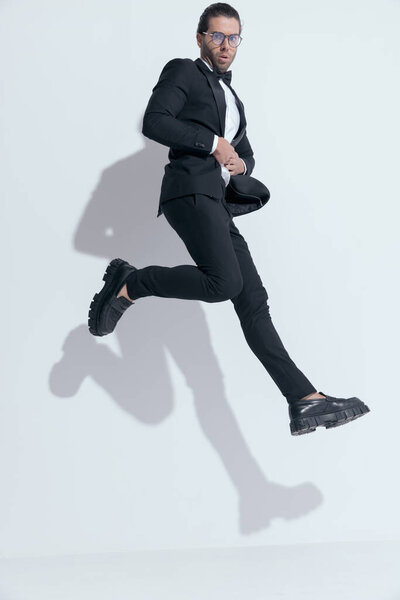 handsome businessman closing his tux and crossing legs in the air, wearing glasses against white studio background