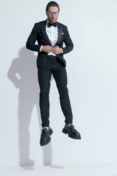 attractive businessman with tough vibe jumping and closing his tux, wearing glasses against white studio background