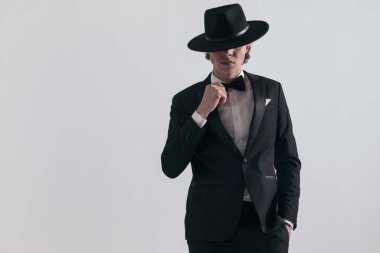 Smokinli gizemli sağdıç papyonu düzeltiyor ve gri arka planda elleri ceplerinde poz verirken şapkayla gözlerini kapatıyor.