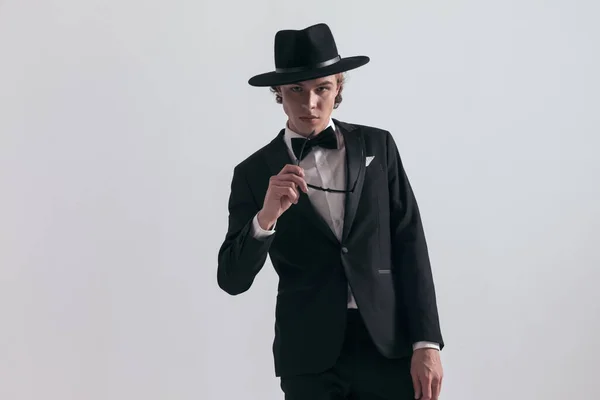 魅力的な男でエレガントなタキシードに帽子を脱いでスタジオで灰色の背景の前でポーズ — ストック写真