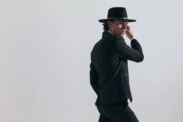 サイドビューのセクシーなエレガントな男で黒タキシードで帽子を探している側とポークで手でポーズ灰色の背景の前 — ストック写真