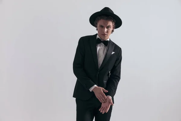 エレガントなセクシー男で黒タキシードで帽子タッチとこすりヤシの前にグレーの背景でスタジオ — ストック写真