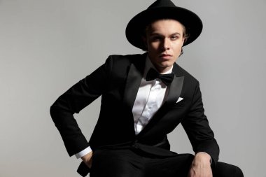 Siyah smokinli zarif genç damat elleri ceplerinde ve siyah arka planda oturuyor.