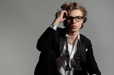 Siyah smokinli zarif bir genç adam. Açık yakalı. Stüdyodaki gri arka planda, yukarı bakıyor ve gözlükleri ayarlıyor.