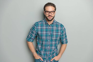 Stüdyoda gri arka planda dururken gözlük takan ve el ele tutuşan ciddi bir gencin portresi.