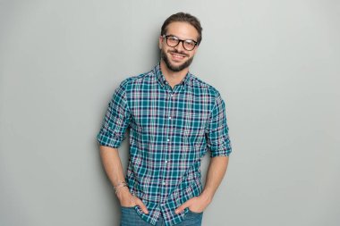 Gözlüklü mutlu bir adam ekose gömlek giyiyor ve ceplerinde el ele tutuşurken stüdyoda gri arka planda poz veriyor ve gülümsüyor.