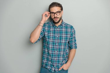 Ekose gömlek ve kot pantolon giyen yakışıklı genç adam gri arka planda el ele poz verirken gözlük ayarlıyor.