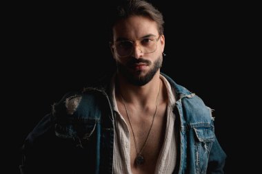 Gözlüklü, kot ceket giyen ve stüdyoda siyah arkaplan önünde poz veren yakışıklı bir gencin portresi.