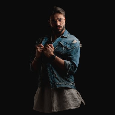 Stüdyoda siyah arka planda ceketini çekerken ve ayarlarken gözlüklü bir moda adamı gözlerini kaçırıyor.