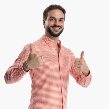 Kırmızı ve beyaz ekose gömlek giyen gururlu genç adam stüdyoda beyaz arkaplan önünde başparmak hareketi yaparken gülümsüyor.