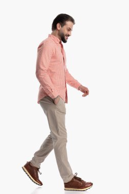 Ekose gömlekli mutlu adamın yan görünüşü. Stüdyoda beyaz arka planda yürürken aşağı bakıyor ve gülüyor.