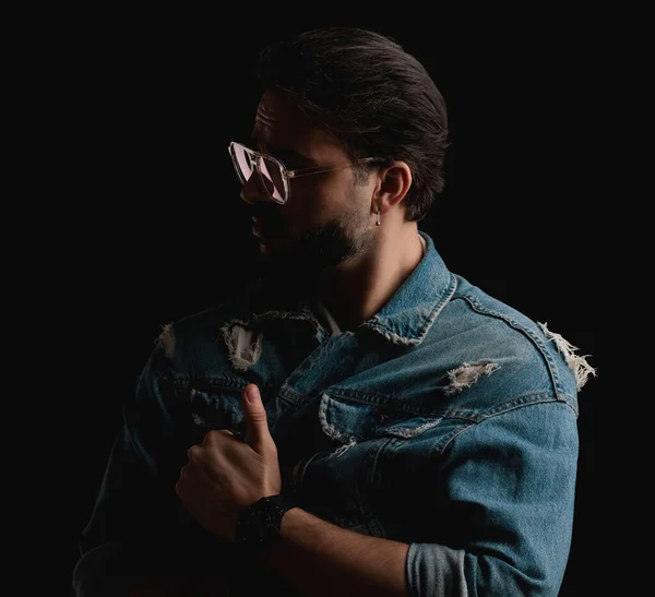 stock image portrait of mysterious young man with glasses closing denim jacket and looking to side in front of black background in studio