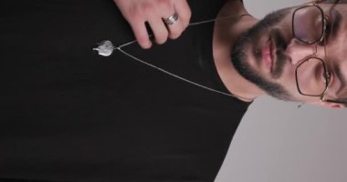 Gözlüklü, avuçlarını ovuşturan, kolyeyi ayarlayan ve gri arka planda sakala dokunurken baştan çıkarıcı olan seksi genç adamın dikey videosu.