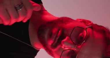 Gözlüklü, avuçlarını ovuşturan ve kolyeyi ayarlamadan önce aşağıya bakan seksi modacının kırmızı ve mavi ışıklarının olduğu dikey video.