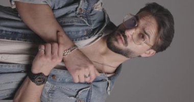 Kot ceket ve gözlük takan seksi moda adamı, giysiyi ayarlıyor ve gri arkaplanın önüne bakıyor.