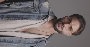 Sıradan bir adam gözlük takıyor, kot ceket tamir ediyor ve düzenliyor, sakala dokunuyor ve gri arka plana bakmadan önce düşünüyor.
