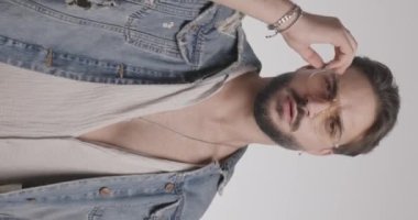 Havalı genç adam göğsünü gösteriyor, kot ceket ve gözlük giyiyor ve ayarlıyor ve açık gri arkaplanın önüne bakıyor.