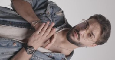 Avuç içlerine dokunan, avuçlarını ovuşturan, mavi kot ceket giyen ve açık gri arka planda ileriye bakan havalı adamın dikey videosu.