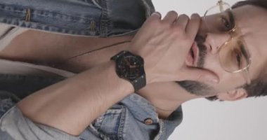 Gözlüklü yakışıklı genç adam açık gri arka planda poz verirken sakal ve mavi kot cekete dokunup düzenliyor.