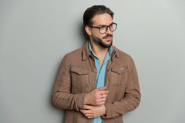 Gözlüklü yakışıklı sakallı adam stüdyoda gri arka planda kollarını düzeltirken yan tarafa bakıyor.