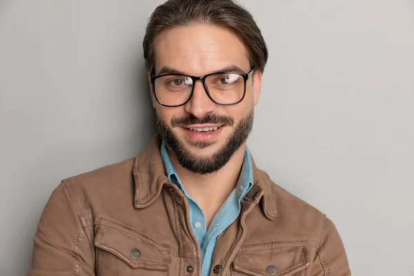 close up portrait of attractive bearded man wearing brown leather jacket and denim shirt underneath smiling and posing in front of grey background