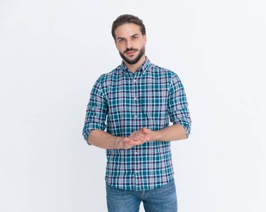 Ekose gömlekli, sakallı, avuçlarını ovuşturan ve gri arka planda kendine güvenen bir şekilde poz veren yakışıklı bir adam.
