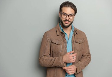 Kahverengi deri ceketli çekici bir adam kollarını düzeltiyor ve gri arka planda poz verirken ileriye bakıyor.